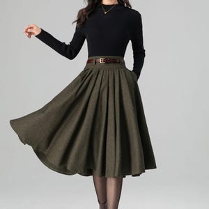 May include: A woman's outfit featuring a black ribbed long-sleeve top and a dark olive green pleated skirt. The skirt is cinched at the waist with a brown belt. The outfit is completed with brown boots and sheer black tights. The overall style is classic and elegant.
