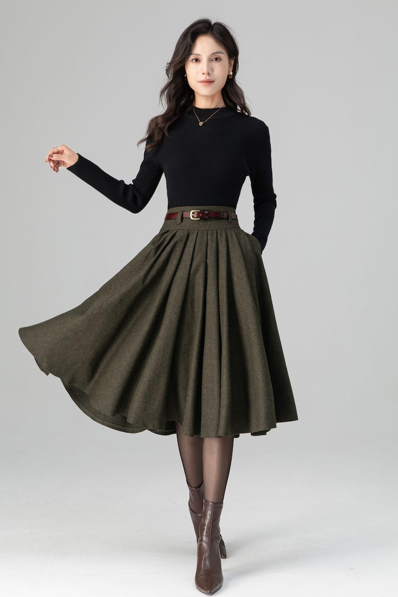 May include: A woman's outfit featuring a black ribbed long-sleeve top and a dark olive green pleated skirt. The skirt is cinched at the waist with a brown belt. The outfit is completed with brown boots and sheer black tights. The overall style is classic and elegant.