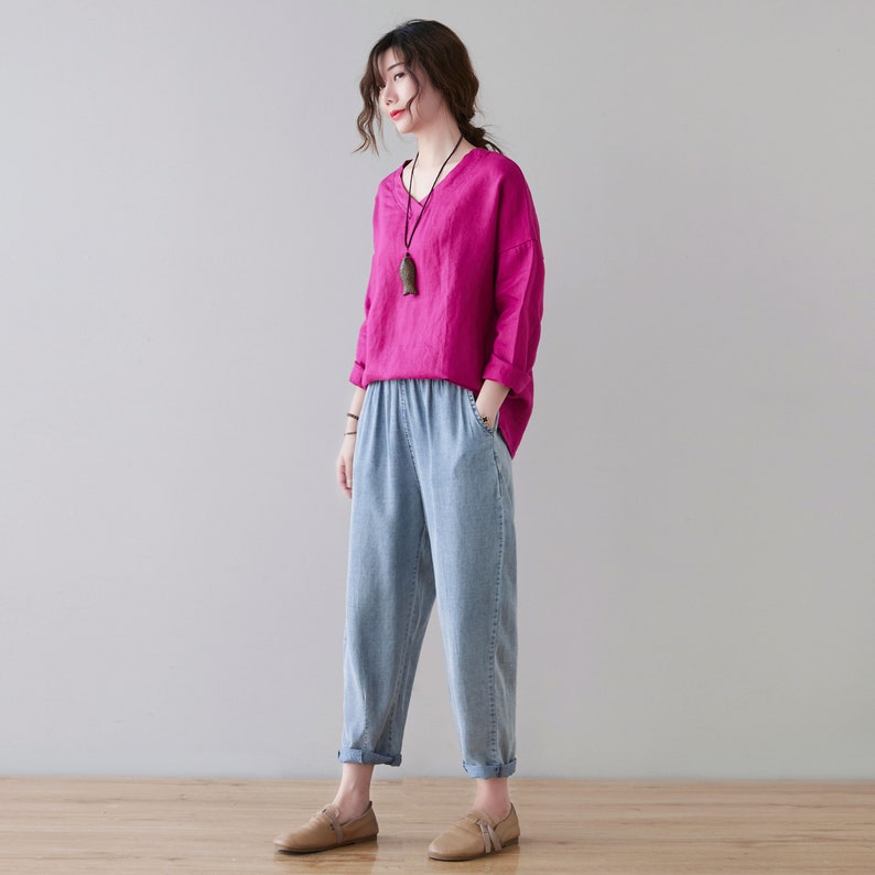 May include: A woman wearing a magenta V-neck long-sleeve shirt and light blue denim pants. The pants have an elastic waistband and are cuffed at the ankles. The woman is also wearing a necklace and brown shoes.