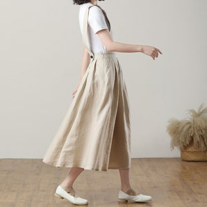 May include: A beige linen overall skirt with a wide, flowing silhouette. The skirt has adjustable straps and a button closure at the waist.