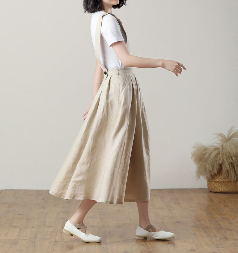 May include: A beige linen overall skirt with a wide, flowing silhouette. The skirt has adjustable straps and a button closure at the waist.