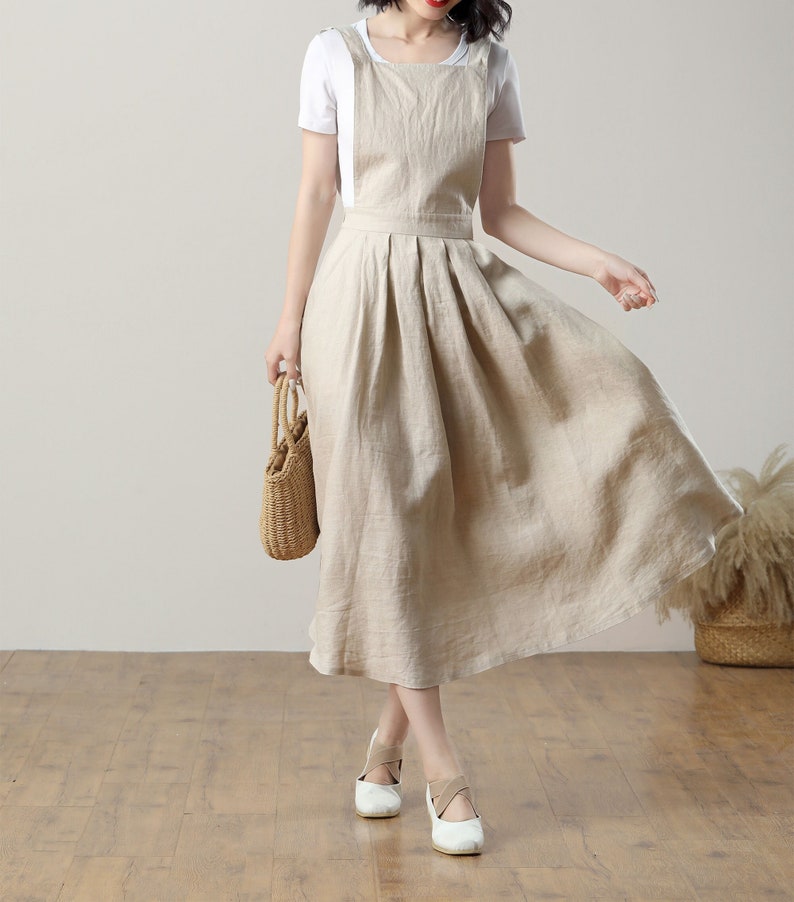 May include: A beige linen overall dress with a white t-shirt underneath. The dress has a square neckline and adjustable straps. The model is wearing white shoes.