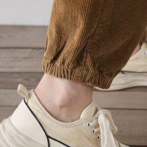 K&ouml;nnte beinhalten: Nahaufnahme des unteren Teils einer braunen Cordhose mit einem wei&szlig;en elastischen Bund. Die Hose wird mit wei&szlig;en Canvas-Sneakern mit schwarzem Besatz getragen.