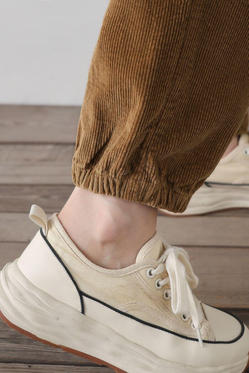 K&ouml;nnte beinhalten: Nahaufnahme des unteren Teils einer braunen Cordhose mit einem wei&szlig;en elastischen Bund. Die Hose wird mit wei&szlig;en Canvas-Sneakern mit schwarzem Besatz getragen.