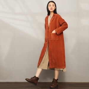 May include: A long, rust-colored corduroy coat with a single patch pocket on the right side. The coat has a notched collar and a button closure.