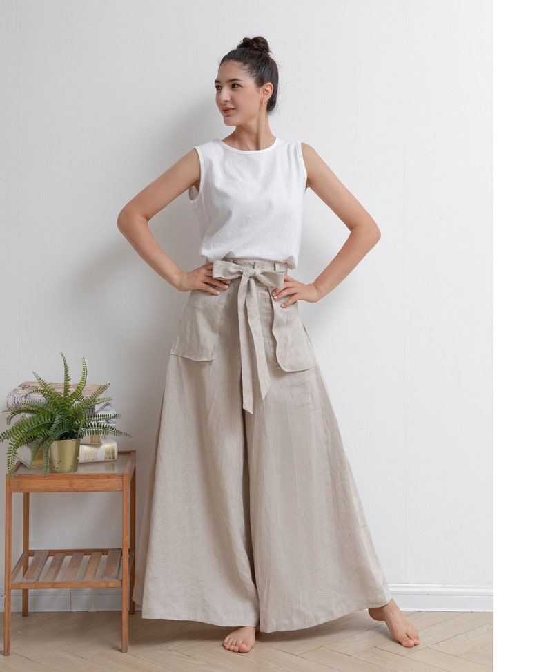 May include: A woman wearing a white sleeveless top and wide-legged linen pants with a self-tie belt. The pants are a light beige color and have pockets on the sides.