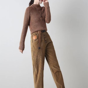 K&ouml;nnte beinhalten: Braune Cordhose mit hoher Taille und Knopfleiste. Die Hose hat einen braunen Lederg&uuml;rtel mit einer Metallschnalle und eine braune aufgesetzte Tasche auf der Vorderseite. Die Hose ist unten umgeschlagen und das Model tr&auml;gt wei&szlig;e Sneaker.
