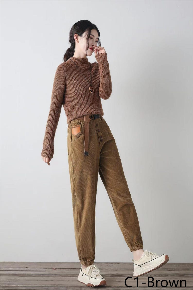 K&ouml;nnte beinhalten: Braune Cordhose mit hoher Taille und Knopfleiste. Die Hose hat einen braunen Lederg&uuml;rtel mit einer Metallschnalle und eine braune aufgesetzte Tasche auf der Vorderseite. Die Hose ist unten umgeschlagen und das Model tr&auml;gt wei&szlig;e Sneaker.