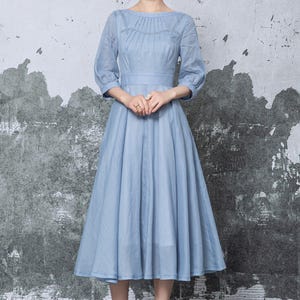 May include: A light blue dress with a round neckline and three-quarter sleeves. The dress features a fitted bodice and a flared skirt, cinched at the waist. The model is wearing white high-heeled sandals. The background is a textured gray wall.