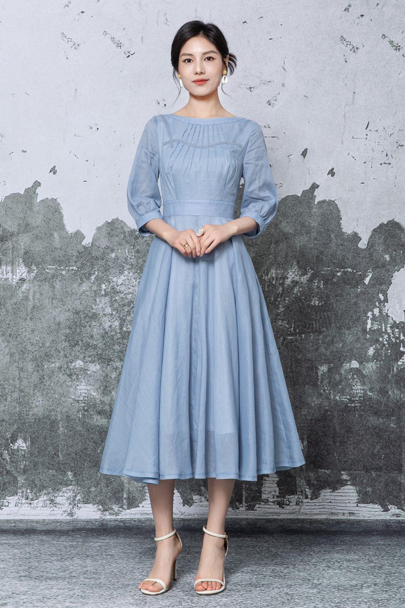 May include: A light blue dress with a round neckline and three-quarter sleeves. The dress features a fitted bodice and a flared skirt, cinched at the waist. The model is wearing white high-heeled sandals. The background is a textured gray wall.