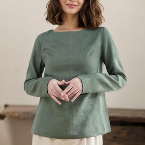 May include: A woman is wearing a long-sleeved, sage green, linen top with a boat neck. She is wearing cream-colored pants.