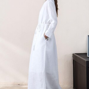 May include: A white long-sleeved maxi dress with a drawstring waist and pockets. The dress is made of a lightweight fabric and has a relaxed fit.