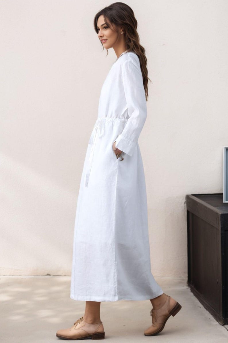 May include: A white long-sleeved maxi dress with a drawstring waist and pockets. The dress is made of a lightweight fabric and has a relaxed fit.