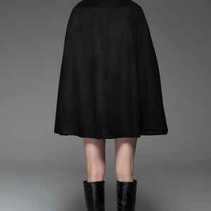 May include: A black wool cape with a long, flowing silhouette. The cape is worn by a person with blonde hair and is styled with black knee-high boots.