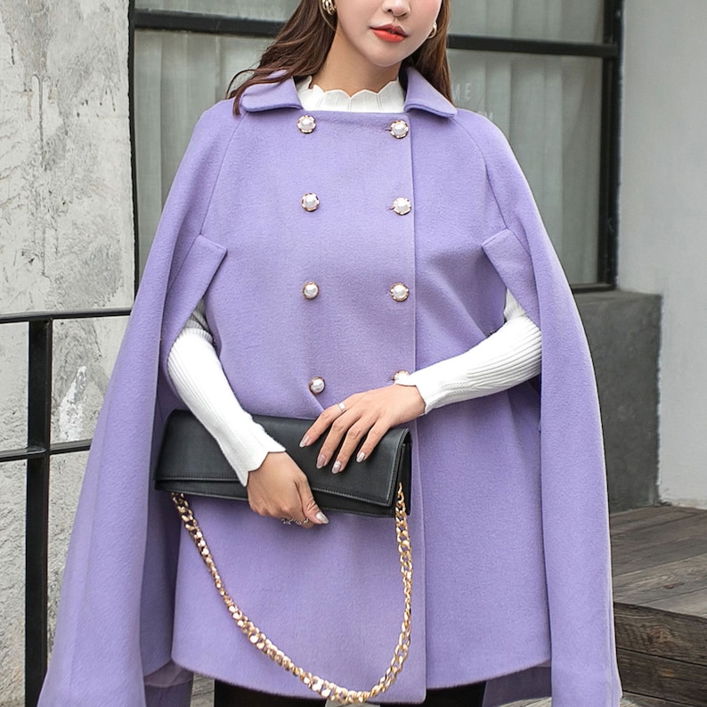 May include: A lavender wool cape with a double breasted closure and pearl buttons. The cape has a chain strap and a black clutch purse.