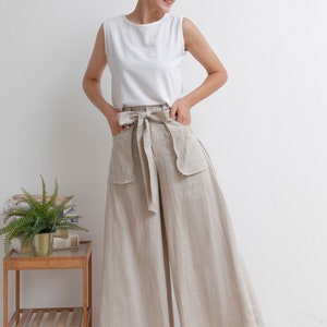 May include: A woman wearing a white sleeveless top and beige linen wide-leg pants with a matching fabric belt. The pants have large patch pockets on the front.