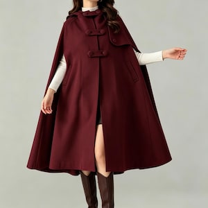 May include: A deep burgundy hooded cape coat featuring a front closure with three horizontal toggle-style buttons. The garment has a smooth texture, a wide hemline, and integrated side pockets, worn over a white long-sleeved top and dark boots.