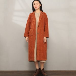 May include: A long, rust-colored corduroy coat with a button closure and two patch pockets. The coat has a relaxed fit and a slightly flared hem.
