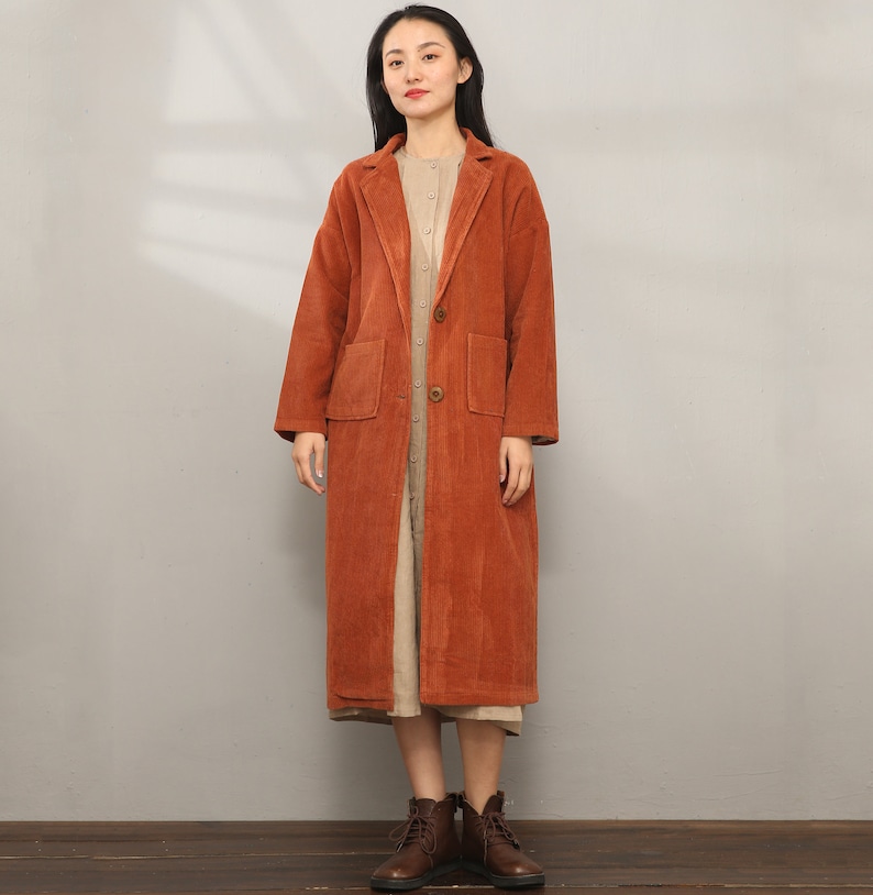 May include: A long, rust-colored corduroy coat with a button closure and two patch pockets. The coat has a relaxed fit and a slightly flared hem.