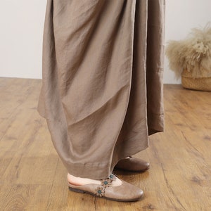May include: A brown linen maxi skirt with a flowing, draped design. The skirt is shown on a model wearing brown leather sandals with floral embellishments.