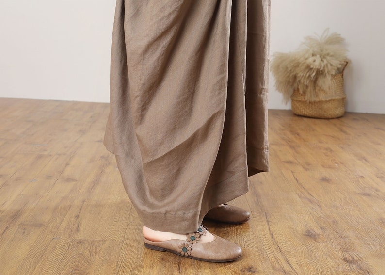 May include: A brown linen maxi skirt with a flowing, draped design. The skirt is shown on a model wearing brown leather sandals with floral embellishments.
