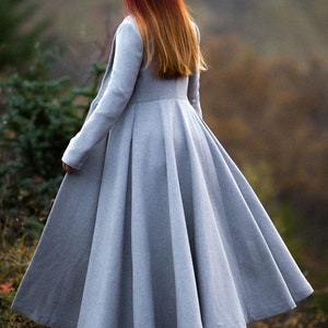 May include: A long, light gray coat with a flared skirt, seen from the back. The coat has a fitted bodice and long sleeves. The person in the coat has red and orange hair. The coat is made of a soft, flowing fabric.