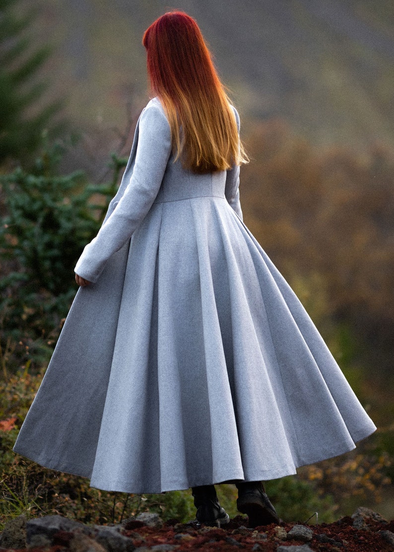 May include: A long, light gray coat with a flared skirt, seen from the back. The coat has a fitted bodice and long sleeves. The person in the coat has red and orange hair. The coat is made of a soft, flowing fabric.