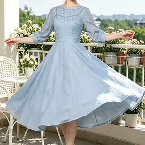 May include: A light blue dress with a flowing skirt and three-quarter sleeves. The dress features a fitted bodice and a defined waist. The model is wearing white high-heeled sandals. The background includes a balcony with white chairs and potted flowers.