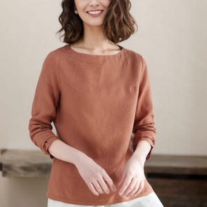 May include: A woman wearing a rust-colored linen top with long sleeves and a boat neck. She is wearing white pants.