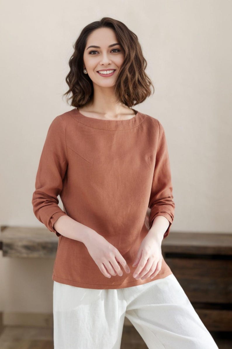 May include: A woman wearing a rust-colored linen top with long sleeves and a boat neck. She is wearing white pants.