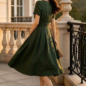 May include: A dark green midi dress with short sleeves, a fitted waist, and a pleated skirt. The dress is paired with a straw hat featuring a black band and tan high-heeled pumps with dark brown pointed toes, while holding a small tan handbag.