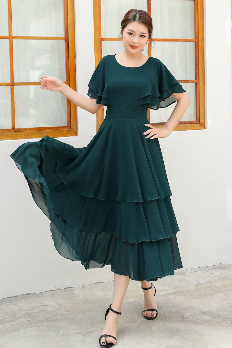 May include: A dark green, tiered midi dress with a round neckline and flutter sleeves. The dress features a fitted waist and a flowing, layered skirt. The model is wearing black sandals.