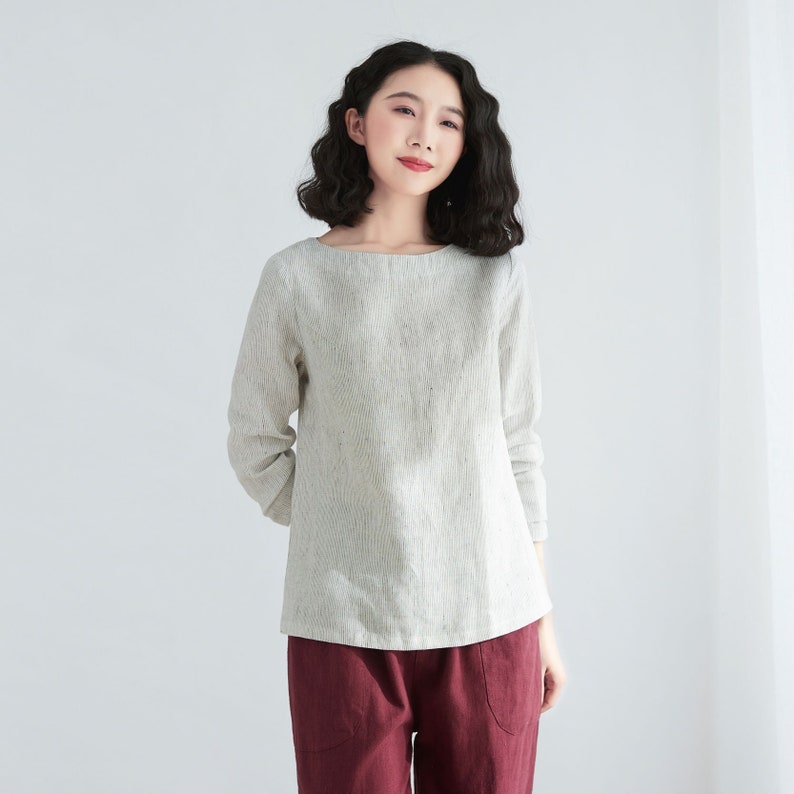 May include: A woman wearing a light gray linen top with a boat neck and long sleeves. She is also wearing maroon pants.