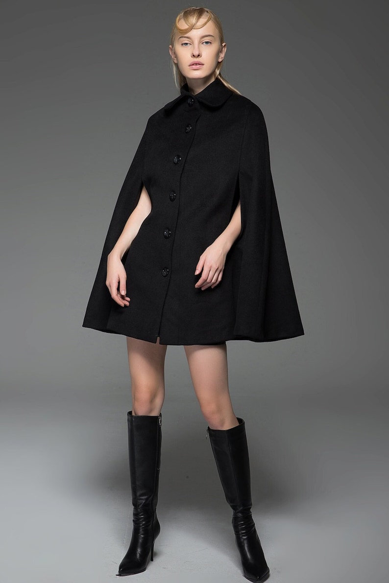 May include: A black wool cape with a button fastening and a collar. The cape is knee-length and has a fitted silhouette. The model is wearing black knee-high boots.