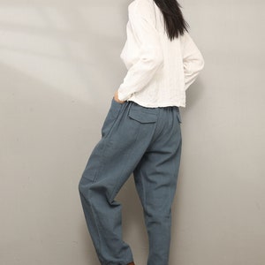 May include: A woman wearing a white long-sleeved shirt and blue linen pants. She is wearing brown ankle boots and has her hair pulled back.
