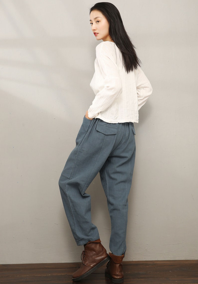 May include: A woman wearing a white long-sleeved shirt and blue linen pants. She is wearing brown ankle boots and has her hair pulled back.