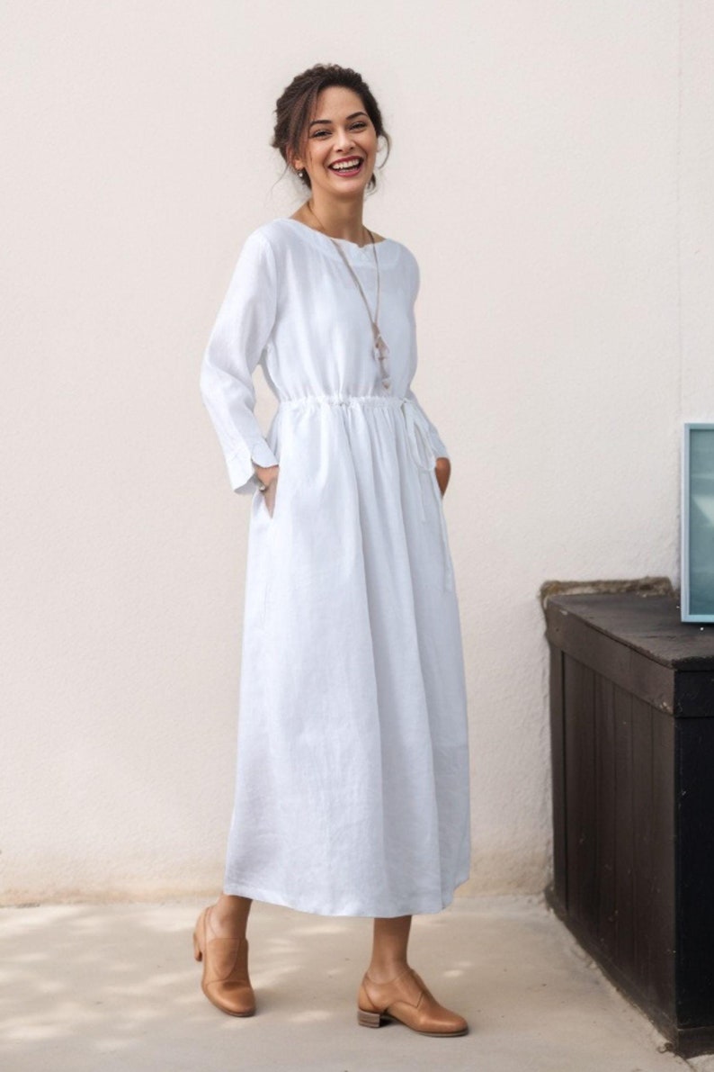 May include: A white linen dress with long sleeves and a drawstring waist. The dress has pockets on both sides and is worn by a person with brown hair and a necklace.
