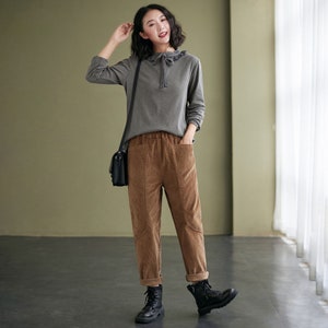 May include: A woman wearing a grey long-sleeved shirt with a ruffled collar and brown corduroy trousers. She is wearing black boots and carrying a black shoulder bag.