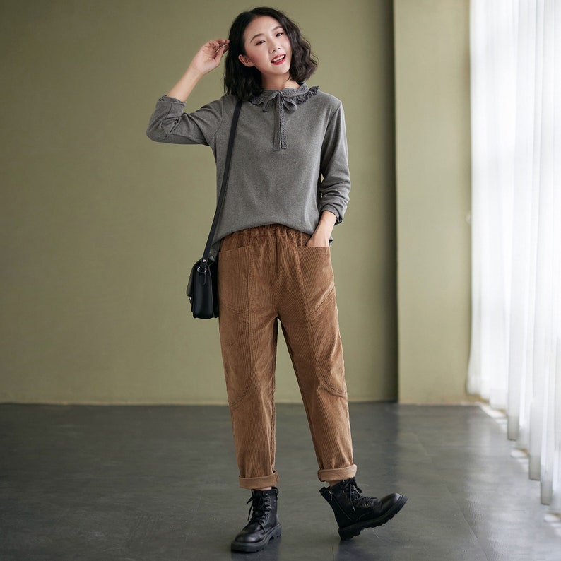 May include: A woman wearing a grey long-sleeved shirt with a ruffled collar and brown corduroy trousers. She is wearing black boots and carrying a black shoulder bag.