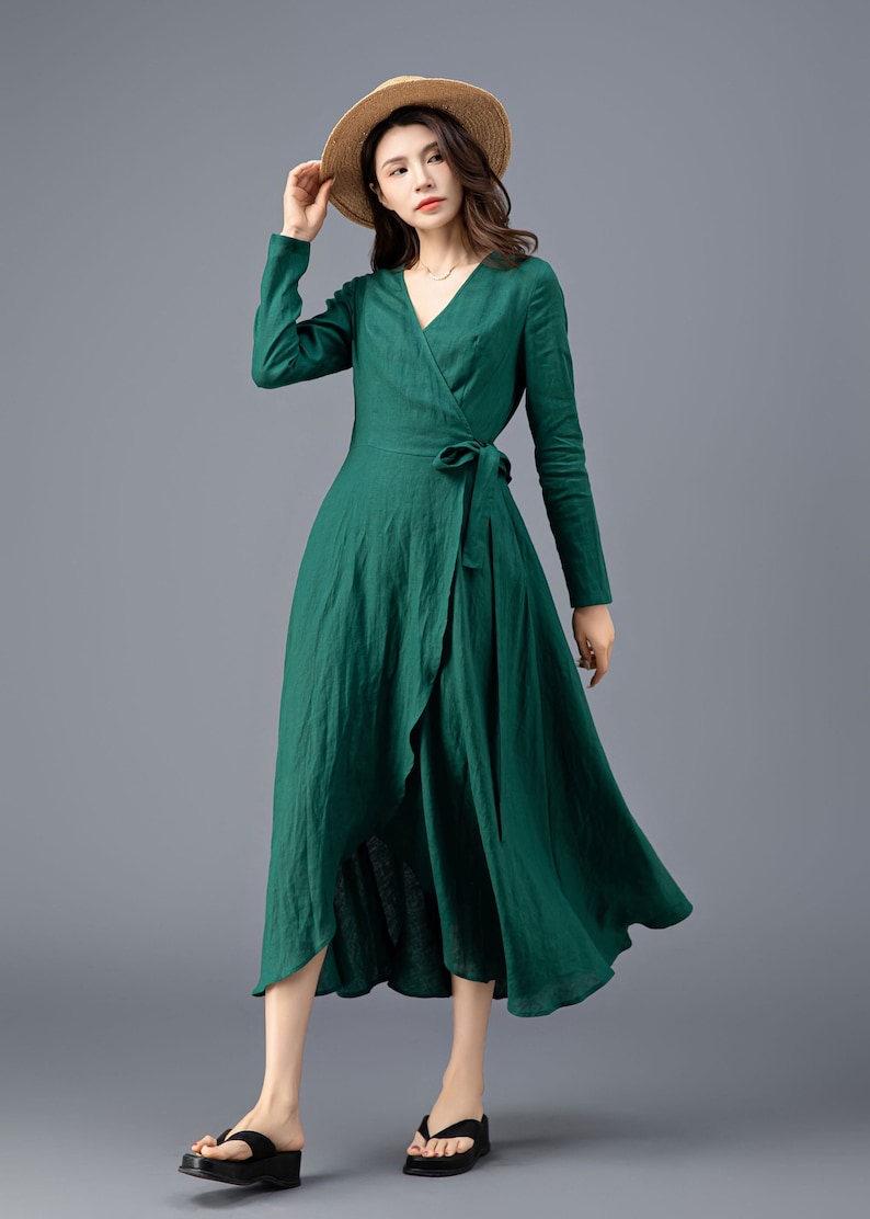 May include: A green wrap dress with long sleeves and a tie at the waist. The dress has a high neckline and a flowing skirt. The model is wearing a straw hat and black sandals.