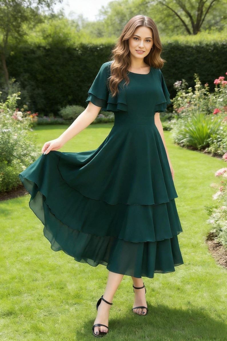 May include: A dark green midi dress with a tiered skirt and flutter sleeves. The dress has a fitted bodice and a flowing skirt. The model is wearing black sandals. The background is a garden with green grass and flowers.