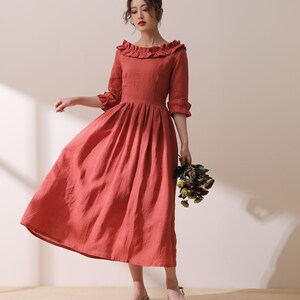 Puede incluir: Un vestido de lino de color terracota con cuello de volantes y mangas 3/4. El vestido tiene un corpi&ntilde;o ajustado y una falda fluida.