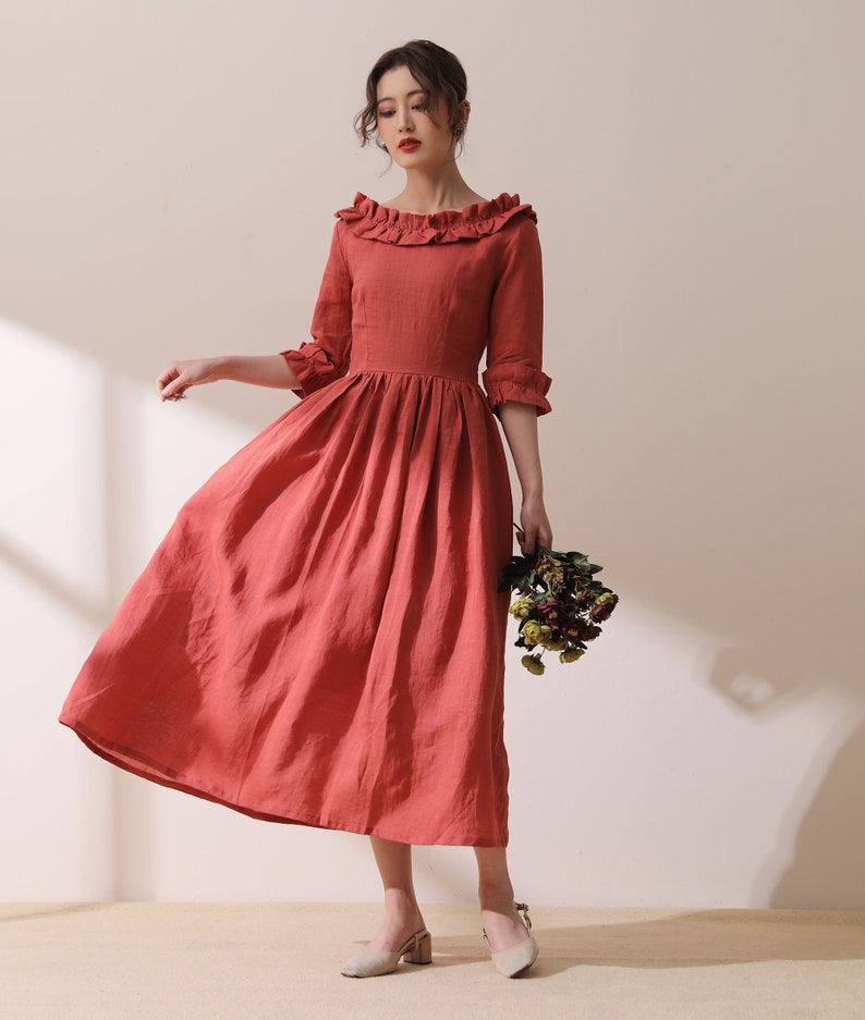 Puede incluir: Un vestido de lino de color terracota con cuello de volantes y mangas 3/4. El vestido tiene un corpi&ntilde;o ajustado y una falda fluida.