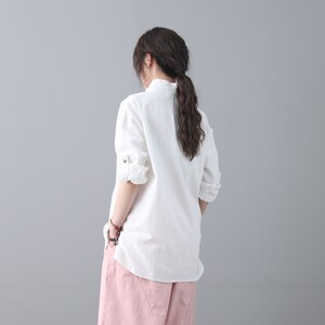 May include: A white linen shirt with a mandarin collar and long sleeves. The shirt is loose-fitting and has a relaxed silhouette. The model is wearing the shirt with a pair of pink pants.