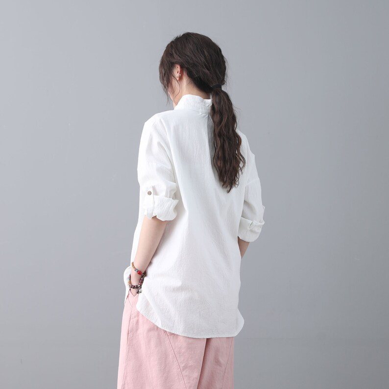 May include: A white linen shirt with a mandarin collar and long sleeves. The shirt is loose-fitting and has a relaxed silhouette. The model is wearing the shirt with a pair of pink pants.