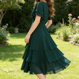 May include: A dark green dress with a tiered skirt and ruffled sleeves. The dress is knee-length and worn with black heels. The woman is standing in a garden with green grass and flowers.