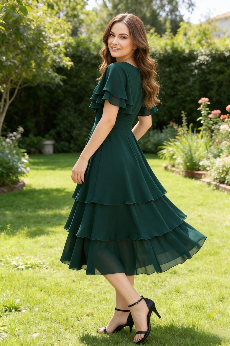 May include: A dark green dress with a tiered skirt and ruffled sleeves. The dress is knee-length and worn with black heels. The woman is standing in a garden with green grass and flowers.