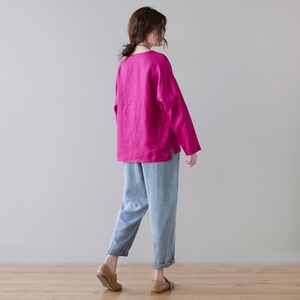 May include: A woman wearing a long-sleeved magenta top and light blue pants. The top has a simple neckline and a relaxed fit. The pants are cuffed at the ankles. The outfit is completed with brown shoes.