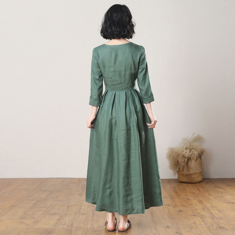 May include: A long, green linen dress with a round neckline and 3/4 sleeves. The dress has a pleated skirt and a fitted bodice.