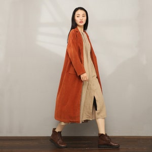 May include: A long, rust-colored corduroy coat with a button closure and pockets. The coat is worn over a beige linen dress.